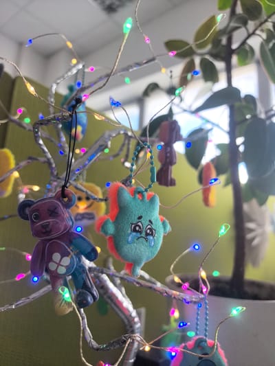 This media file shows a close-up view of a decorative tree adorned with small, colorful trinkets and LED fairy lights. The subjects are primarily objects: the trinkets appear to be small plush toys or charms, including a purple bear with an X for an eye and a blue creature with a sad expression, both hanging from the tree branches. The tree itself is decorated with metallic, foil-wrapped branches. The fairy lights are small, multi-colored LEDs that are lit, creating a festive and whimsical atmosphere. In the background, a portion of a green plant in a pot is visible, along with a blurry green wall and a bright window, suggesting an indoor setting.
There is no apparent activity or event taking place, as the scene appears to be a still, decorative arrangement. Notable details include the variety of light colors (blue, green, pink, yellow, orange) and the textures of the trinkets and the tree branches. The overall mood is cheerful and playful due to the lights and colorful decorations, despite the sad expression on one of the trinkets. The time of day is not clearly indicated, but the bright background suggests it might be daytime. Location cues such as the green wall and plant are common in indoor environments. No visible text is present in the image.
