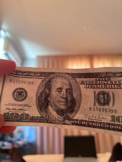 The image depicts a partially visible, vertical, and slightly crumpled United States one hundred dollar bill. The bill is held by a person out of frame. The serial number B17696399 is visible in two locations. The signature of the Secretary of the Treasury, Robert E. Rubin, can be seen. The portrait of Benjamin Franklin is centrally located on the bill. The background is blurred, showing a light-colored wall on the left and part of what appears to be a room with blinds or a patterned surface on the right. The location in the city of Kribul, Bulgaria, cannot be determined from the visual information provided.
