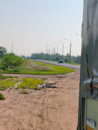 The image captures a roadside scene near Krasnokholm, Russia. In the foreground, a dirt path shows tire tracks and sparse vegetation. To the right, a portion of a large vehicle, possibly a truck, is visible, featuring a gray tarp and a white panel with the partial inscription "CHMITZ". Beyond the path, a paved road stretches into the distance, lined with a row of streetlights. A black car is traveling on the road. Green grass and trees flank the road, with a tall, slender structure, possibly a radio tower, visible in the background on the left. The sky is clear and pale blue.