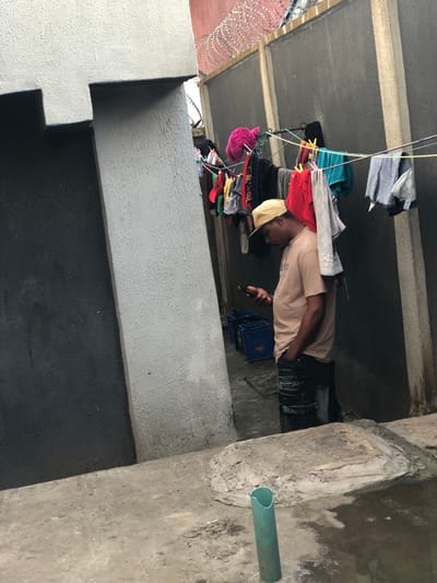 An outdoor, somewhat cramped space is depicted. Clothing hangs on lines, suggesting laundry. A person wearing a light-colored shirt and hat is visible, possibly using a mobile phone. A blue crate is near the individual. The concrete structure surrounds them. A green pipe protrudes from a wall. The walls are layered with black and gray paint. Barbed wire tops a wall section.