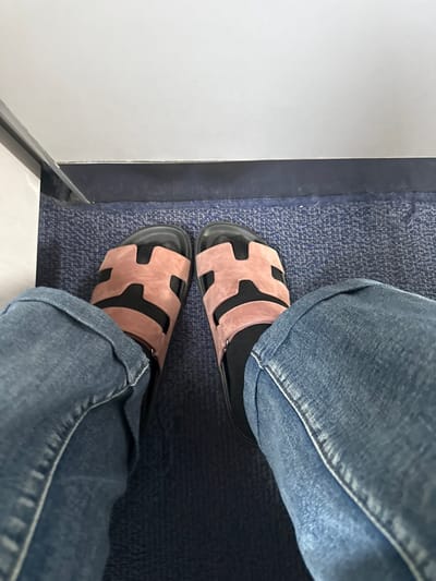 The image depicts a first-person perspective of an individual's lower legs and feet, located in Khoronq, Armenia. The subject is wearing blue denim jeans with visible cuffs and black socks. On their feet are a pair of light pink open-toed sandals featuring black soles and a distinctive H-shaped cutout design on the upper straps. The feet rest on a blue, mottled, low-pile carpet. In the background, a dark blue or black baseboard separates the carpet from a plain white wall or partition above it. The scene is static, showing the person's feet and the immediate environment.