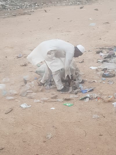 A man wearing a white cap and white clothing is kneeling amidst scattered trash and debris on a dirt or sand surface. The debris includes plastic bags and wrappers. The location is Dutsinma A, Nigeria.