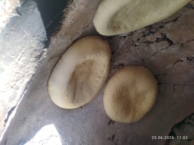 Three flatbreads are shown, two on a rough, cracked, reddish-brown surface and one partially visible above them. The bread appears to be lightly browned and has a textured surface, possibly from the baking process or from being pressed. The lighting suggests an outdoor or partially enclosed environment, with shadows cast on the surface. The timestamp "25.06.2026 11:02" is visible in the lower right corner. The bread is positioned inside what appears to be a traditional Kyrgyz oven, possibly a tandoor, judging by the curved, rough interior walls and the context of traditional breadmaking in Kyrgyzstan. The location in Özgön, Kyrgyzstan, suggests this is a scene of local food preparation.