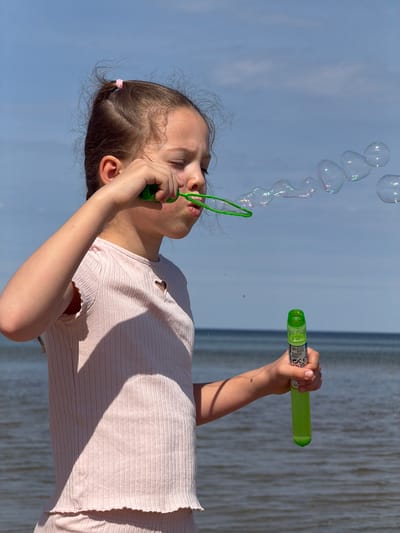 A young girl stands against a backdrop of a light blue sky and a body of water. She is blowing bubbles with a green bubble wand, creating a chain of iridescent spheres in the air. In her right hand, she holds a green bubble solution container. She is wearing a light pink ribbed shirt. The location is likely near a body of water in Plieņciems, Latvia, judging from the water and sky conditions. The girl's expression is focused as she engages in the activity.