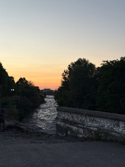 The image depicts a canal at sunset. The sky displays a gradient of pale blue to warm orange, indicating the end of the day. Lush green trees line the banks of the canal, their dark silhouettes contrasting with the vibrant sky. The water's surface reflects the colors of the sunset, creating a shimmering effect. A weathered stone bridge or embankment runs along the right side of the canal, its textured surface showing signs of age. Beyond the trees, in the distance, buildings and streetlights are faintly visible, suggesting an urban or semi-urban setting near the canal. The overall mood is tranquil and serene, characteristic of a sunset by the water. The time of day is clearly sunset. No people or specific text are visible in the image.