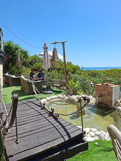 A man and a woman are seated at a table on a wooden deck overlooking a small pond. The woman is wearing a black t-shirt and the man is wearing a dark green t-shirt. They are both looking down at something on the table. The scene is set outdoors with lush green vegetation and the blue ocean in the background, suggesting a coastal location like La Mata, Spain. The sky is a clear, bright blue, indicating a sunny day. Two umbrellas are partially visible, suggesting the area is set up for outdoor dining or relaxation. The pond has a stone border and a small brick structure, possibly a decorative fountain, with water flowing into it. A wooden walkway or deck surrounds part of the pond. The overall atmosphere appears tranquil and leisurely. There is no visible text in the image.
