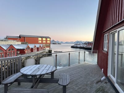 The image displays a waterfront scene in Stamsund, Norway, observed from a wooden deck during twilight hours. In the foreground, a weathered wooden deck surface is visible, bearing traces of frost or moisture. A circular wooden picnic table with integrated benches, also lightly frosted, is positioned on the deck. A contemporary railing system, consisting of clear glass panels secured by metal posts, defines the deck's perimeter. To the immediate right, a portion of a red-painted wooden building with vertical siding and white-framed windows is present.
Across the calm water, a complex of red-painted wooden structures is observed. This includes a row of smaller, gabled cabins (rorbuer) with snow-dusted roofs, connected by a wooden walkway or pier. Behind these, a larger, multi-story red building with a dark roof and illuminated windows is situated. Further in the distance, another red building is visible on a snowy point of land. The background features a range of rugged, snow-capped mountains under a gradient sky that transitions from light blue overhead to a pale orange-pink near the horizon. No individuals are depicted in the scene.