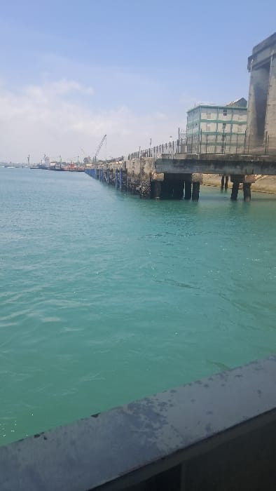 The image depicts a daytime view of a marine industrial environment identified as the Ganjoni/Shimanzi ward in Kenya. In the immediate foreground, a weathered, dark grey concrete or metal railing is partially visible, extending horizontally across the lower right. The midground is dominated by a body of turquoise-green water, exhibiting minor surface ripples and light reflections. To the right, an aged concrete pier or dock structure, supported by multiple concrete pillars, extends into the water. This structure shows signs of material degradation and biological growth on its submerged sections. A black metal fence runs along the top of the pier. Further along the pier, a multi-story building with a light green facade and white window frames is situated on the landward side. In the background, along the distant coastline, multiple large cargo ships, a prominent lattice boom crane, and various other port infrastructure elements are discernible, indicating commercial shipping operations. The sky above is clear and light blue with sparse white cloud formations. No individuals, explicit actions, or direct interactions are visible. The overall scene is brightly illuminated by natural light.