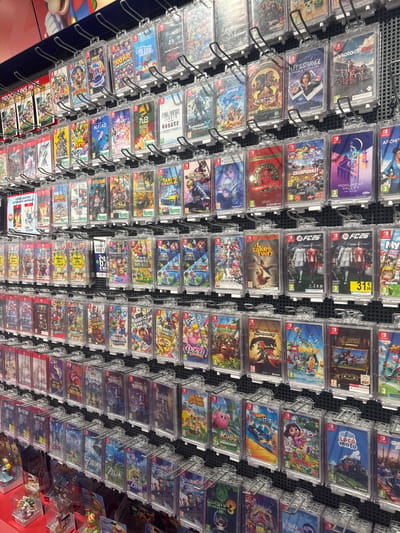 The image displays a retail display of video games, primarily Nintendo Switch games, in a store located in Leers, France. The games are organized on shelves and hung from hooks, each encased in a clear plastic protector. The titles visible include popular franchises such as "The Legend of Zelda," "Super Mario," "Pokémon," "Final Fantasy," and various sports games like "MotoGP." Some games have price tags or age rating indicators. The store environment appears to be well-lit, with a focus on showcasing the merchandise. There are no people clearly visible in the frame, and the focus is solely on the product display. The background suggests a typical retail setting with shelving units and possibly other store fixtures.