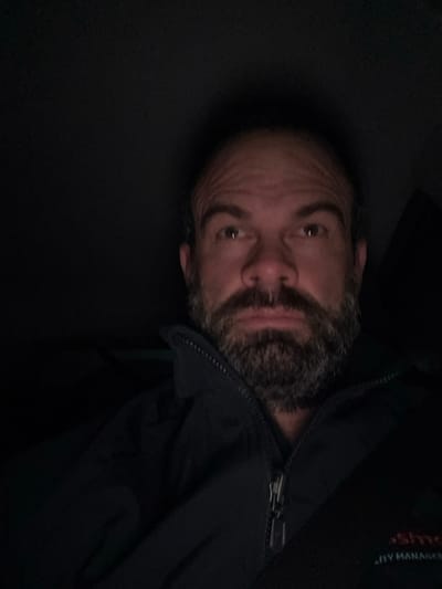 A man with a salt-and-pepper beard and a partially shaved head is angled to the right in dim lighting. He's wearing a dark jacket, with a visible zipper. His eyes are directed towards the viewer. The background is mostly dark and undefined, with hints of a darker fabric or surface. The overall image appears underexposed.