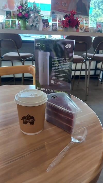 The scene depicts a table with a coffee cup, cake, and a spoon from the Punthai coffee shop in Plai Phraya, Thailand. A menu is visible standing on the table with details about the Punthai GenZero program that encourages customers to bring their own reusable cups for a discount. Chairs are visible behind the table and flowers can be seen in the background.