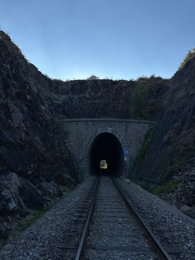 The image shows a railway tunnel entrance surrounded by steep, rocky terrain. The tunnel has an arched opening constructed with stone blocks, and train tracks extend from the foreground into the darkness of the tunnel. The sky above is a clear, pale blue. There are no people or animals visible in the image. The environment appears natural and rugged, with some vegetation growing on the slopes. A few signs are visible on the tunnel's exterior wall. The scene suggests an isolated and somewhat remote location. Based on the provided context, this is the entrance to the train tunnel in Générargues, France.
