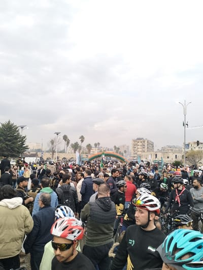 A large crowd of people gathers in a public square, some wearing helmets and seemingly participating in a cycling event. An inflatable archway is visible in the background, possibly marking the start or finish line. The crowd consists mostly of men, and some are holding bicycles. The sky is overcast, and buildings and trees can be seen in the surrounding area. The overall atmosphere suggests a community event or public gathering.
