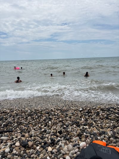 This image captures a scene at the pebble beach of Psyrdzkha, Abkhazia. The sky is overcast with scattered clouds. Several people are visible in the sea, engaged in swimming and recreation. In the foreground, a collection of various sized pebbles covers the beach. In the middle ground, the water is moderately choppy with small waves breaking near the shore. Several individuals are dispersed in the water, some appearing to be children. One person is near a pink inflatable ring. In the background, the sea extends to the horizon, meeting the cloudy sky. Two life vests are visible on the pebble beach in the lower right corner of the image.