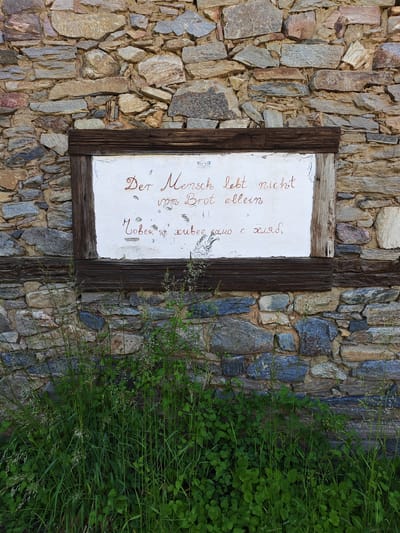 This is a medium shot of a section of a rustic stone wall in Kovachevitsa, Bulgaria, featuring a framed plaque and dense undergrowth. The wall is constructed from irregularly shaped, multi-toned natural stones, predominantly grey, beige, and brown, with visible mortar lines. A rectangular plaque, framed by dark, distressed timber, is recessed into the upper-middle portion of the wall. The plaque surface is white and shows signs of weathering, with minor paint chips and texture variations.
Written on the plaque in a reddish-brown, cursive script are two lines of German text: "Der Mensch lebt nicht" followed by "vom Brot allein" (Man does not live by bread alone). Below the German text, a line in Cyrillic script reads: "Човек не живее само с хляб" (Man does not live by bread alone), which is the Bulgarian translation. Small, faint decorative motifs are present in the upper corners of the white plaque.
The lower half of the image is dominated by lush green vegetation, including tall grasses with developing seed heads and various broad-leaved plants, growing densely from the ground against the base of the stone wall and partially obscuring the bottom section of the wooden frame. The scene is static, captured under natural daylight, emphasizing the textures of the stone, wood, and plant life. There are no people or specific actions depicted.