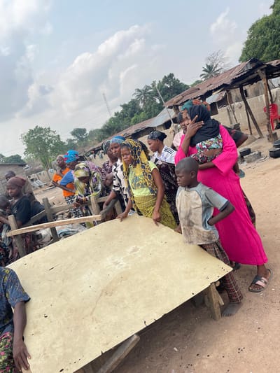 An outdoor scene in Osara Gada, Nigeria, depicts approximately a dozen individuals, predominantly women and children, gathered around a large, light-colored, rectangular wooden or composite board serving as a makeshift table. The table, positioned horizontally in the foreground-midground, rests on rudimentary wooden supports and exhibits surface wear and imperfections.
The ground is unpaved, consisting of light brown dirt. The sky is overcast with visible white and grey clouds. In the background, several single-story structures with corrugated metal roofs are visible, alongside green foliage, palm trees, and a distant metal tower. An outdoor cooking area with pots and a red plastic chair is visible to the far right.
Individuals present include: a young boy in a stained light blue shirt and plaid pants, standing barefoot next to the table on the right; a woman in a bright pink dress holding an infant wrapped in a patterned cloth, with the infant's head covered by a black scarf; and multiple adult women, most wearing headscarves and patterned traditional garments. Some women are leaning on the makeshift table, while others stand behind it or observe from various positions. One woman wears a prominent yellow and black patterned headscarf and a green top. Another woman wears an orange top and a blue headscarf further to the left. No explicit actions or interactions beyond gathering around the table are discernible.