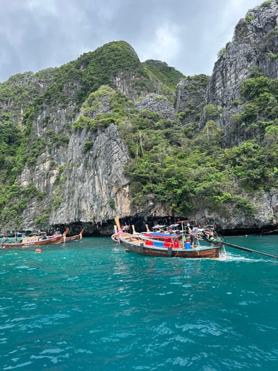 The image captures a scenic view of turquoise waters with several longtail boats navigating near a towering limestone cliff face covered in lush green vegetation. The cliff is rugged and appears to be part of a coastal landscape. The boats are traditional Thai longtail boats, characterized by their wooden hulls and long poles with propellers. One boat in the foreground has people on board, with one individual operating what appears to be the engine. The water is clear and reflects the overcast sky. The scene suggests a tropical location, likely a bay or cove, common in areas like Haad Yao, Thailand, known for its karst topography and beautiful seascapes.