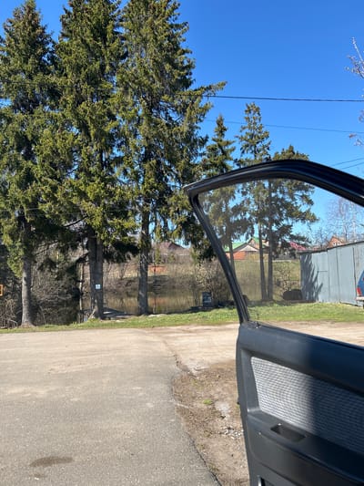 The image depicts a view from inside a car with an open door, showing an exterior scene that features a paved road, bordered by grassy areas and a body of water. In the foreground are tall evergreen trees, which partially obscure the view of houses and buildings in the background. Overhead power lines are visible against a clear blue sky. The interior of the car is seen through the open door, showcasing a gray door panel. The location is specified as Bashur, Russia.