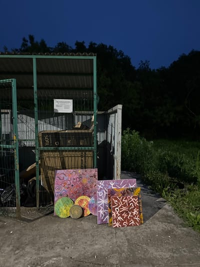An outdoor scene at dusk in Vilyuchinsk, Russia, features a green metal dumpster enclosure with corrugated metal roofing. Inside the enclosure, several cardboard boxes are visible, including one labeled "YAOS" and another with Cyrillic text indicating "ВИЗОР-LED." To the right of the dumpster, a collection of paintings is arranged on the ground. These include a large rectangular painting with swirling pink and orange abstract designs, three circular paintings with green, yellow, and red hues, and two smaller square paintings with geometric patterns in purple and brown. The surrounding environment consists of a concrete surface, tall green grass, and dense trees in the background, all illuminated under a deep blue twilight sky.