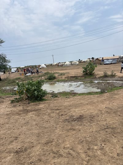 The image depicts a scene in the city of Ad-Damazin, Sudan. In the foreground, a muddy puddle sits on dry, dusty ground with scattered debris. To the left, a green bush is prominent. In the middle ground, a group of approximately ten individuals are gathered around a makeshift structure with a thatched roof. Several white tents and some more rudimentary shelters are visible in the background, spread across a gently sloping, barren landscape. Power lines are strung across the upper portion of the image, and a single utility pole stands to the right. The sky is overcast with clouds.