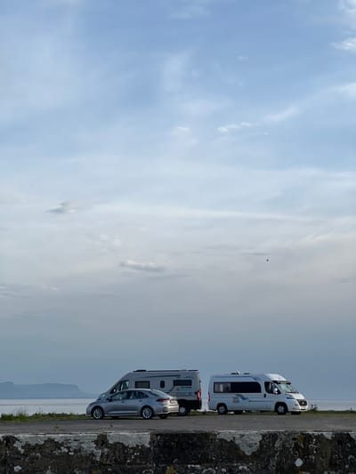 This image depicts a scenic view in Terhillion, Ireland, featuring a coastal landscape under a partly cloudy sky. In the foreground, a low stone wall with a textured surface runs horizontally across the bottom of the frame. Behind the wall, a paved area is visible, where two vehicles are parked. To the left, a silver sedan is positioned next to a grey camper van. To the right of the sedan and camper van, a white camper van is parked. The vehicles are facing towards the right of the image. In the background, a body of water stretches towards the horizon, with a distant landmass or mountains silhouetted against the soft, overcast sky. The overall impression is one of tranquility and a picturesque natural setting.