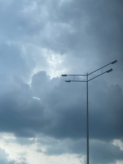 A low-angle shot captures a sky dominated by thick, grey storm clouds. In the upper portion of the frame, a metal utility pole extends horizontally. Attached to the pole is a vertical arm supporting two streetlights. Isakovo, Russia.