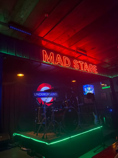 The image depicts the interior of Mad Stage, a music venue located in Tampico, Mexico. The scene is dimly lit with a combination of neon and ambient lighting.

A prominent red neon sign spelling "MAD STAGE" is mounted on a textured red and brown wall. Adjacent to the sign is a London Underground logo illuminated with blue and red neon. A drum set is visible behind the London Underground sign.

On the other side of the drums are a green neon light, and a beer sign. A television is showing a video of some type. A black bass guitar is behind the drum set. The walls are covered in brown and green wood paneling.