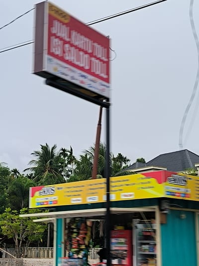This image captures a blurred street scene in Glee Gogo, Indonesia, appearing to be taken from a moving vehicle under an overcast daytime sky.
A prominent red and white billboard, heavily motion-blurred, dominates the upper left. Despite the blur, the text "JUAL KARTU TOLL ISI SALDO TOLL" is discernible, indicating that the establishment sells toll cards and offers toll balance top-up services. This billboard is supported by a dark utility pole.
Below, a small kiosk or storefront is visible, featuring a bright yellow and red awning sign. On the red part, the word "GENIUS" is somewhat clear, and on the yellow section, a logo resembling "SHOPEEPAY" along with other blurred service icons can be identified, suggesting mobile payment or digital service offerings. The kiosk itself has a distinct teal or turquoise-colored wall and contains what appear to be display refrigerators filled with items, though these are also heavily blurred.
The background consists of dense green foliage, predominantly palm trees, characteristic of a tropical setting. Behind the trees on the right, the dark tiled roof of a house is visible. A thin black cable stretches across the upper left portion of the frame. The scene conveys a commercial area along a road, bustling with activity despite the motion blur.