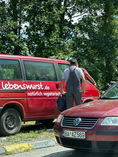 In the city of Dulovine, Montenegro, two individuals, presumably workers, are near a red van with the text "Lebenswurst.de" and "natürlich vegetarisch" on its side. One person, wearing grey overalls over a grey t-shirt, is facing away from the camera and appears to be either interacting with the van or carrying a dark jacket. Another person, wearing a red t-shirt, is partially visible behind the first individual, reaching towards the van. A dark red car is parked in front of the van, with its license plate reading "BA AZ 569". The vehicles are on a paved surface next to a curb with yellow and grey markings. Green foliage and trees are visible in the background, suggesting an outdoor, possibly park-like or roadside setting.
