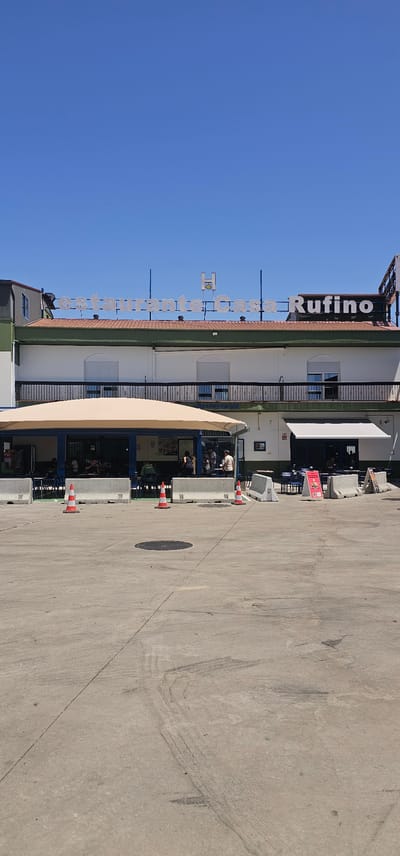 The image captures a building with a sign that reads "restaurante Casa Rufino" in Aldea del Cano, Spain. The building has a white facade with green trim and a tiled roof. In front of the building is an outdoor seating area covered by a beige awning. Several tables and chairs are visible, and some people are seated, suggesting it is open for business. The foreground shows a concrete area with three orange and white traffic cones and some concrete barriers. The sky is clear and blue, indicating a sunny day. The overall scene depicts a restaurant exterior in a somewhat open, possibly commercial or recreational area.
