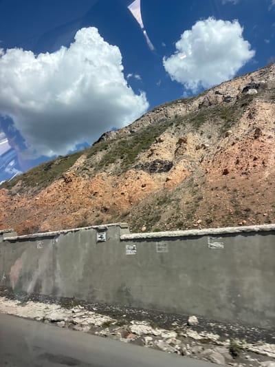 The image shows a steep, rocky mountainside in Nurnus, Armenia. Patches of vegetation cling to the rock face. Adjacent to the mountainside is a gray concrete structure, possibly a retaining wall or a section of infrastructure. Above, a blue sky is dotted with white, fluffy clouds. The viewpoint suggests the image was taken from an elevated position, potentially from within a vehicle. The overall impression is a landscape where human construction meets natural geological features.