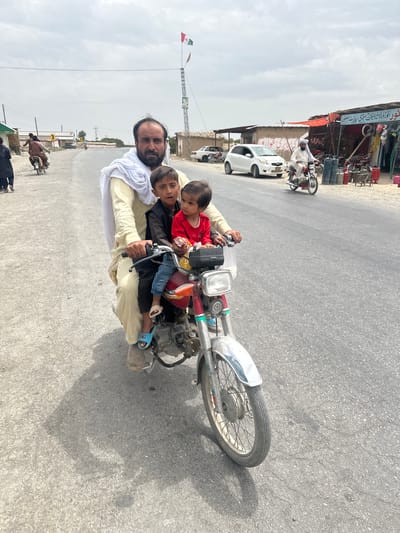 In Kan Mehtarzai, Pakistan, a man with a beard and a white shawl draped over his shoulder rides a motorcycle. He is carrying two young children on the motorcycle. The child in front is wearing a black shirt, and the child behind is wearing a red shirt. The motorcycle is red and silver. In the background, there are buildings, a white car, and a flag on a pole. The sky is cloudy.