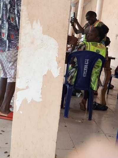 The image depicts an indoor or covered area in Ajalli, Nigeria, likely a public or administrative setting. In the foreground, a prominent beige structural column or wall section shows significant white paint or plaster peeling. To the immediate left of this column, a person's lower legs are visible, wearing white patterned shorts with a repeating dark logo, a dark patterned shirt, and orange slip-on sandals.
To the right of the foreground column, a female individual is seated, facing right, on a blue plastic chair. She wears a bright yellow-green reflective safety vest over a patterned dress. The vest displays text, including "PO" and "PRESIDING OFFICER 1" on the back. Her dark hair is pulled back into an updo. The blue plastic chair has "BB RENTALS" embossed vertically on its backrest. Behind her, a male individual stands, holding a dark-colored smartphone horizontally at eye level, seemingly capturing an image or video to his right. He also wears a reflective vest.
The background consists of light-colored walls and partial views of classical-style architectural columns behind the standing male. A wooden table or desk is partially visible behind the seated woman. The floor is covered with light-toned square tiles and shows scattered dark debris, possibly dirt or small stones, along with two black plastic bags situated near the chairs.