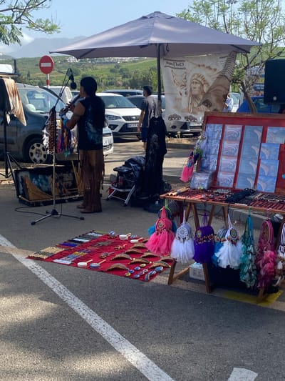 The image depicts an outdoor market scene in San Luis De Sabinillas, Spain, under a clear blue sky. A person dressed in brown fringed pants and a dark t-shirt with a wolf graphic is playing a flute. In front of the musician, a red cloth is spread on the ground, displaying an assortment of beaded necklaces, bracelets, and small decorative items. To the right, a wooden stall is set up, showcasing CDs with Native American-themed artwork and various handcrafted items, including dreamcatchers with colorful feathers hanging from the stall. Behind the stall, a large umbrella shades the area, and a tapestry with an image of a Native American chief and a wolf is displayed. Several cars are parked in the background, suggesting a roadside market or parking lot. The environment appears to be a coastal area with hills and greenery visible in the distance.
