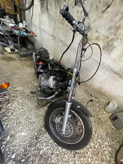 An indoor scene shows a partially disassembled motorcycle, likely a cruiser or chopper, lying on its left side on a garage floor. The motorcycle features a large, chromed V-twin engine with visible cooling fins and exhaust pipes. Its front end includes a long, chromed fork assembly, a front wheel with a disc brake and spoked rim, and handlebars equipped with grips and brake/clutch levers. Various colored wires (red, green, yellow, blue, black) are exposed around the steering head and engine, suggesting ongoing repair or customization work.
The floor beneath the motorcycle is a mix of rough concrete and light-colored gravel, with a beige tarp or fabric laid out under the engine area. In the background, against a rough concrete wall, several items are visible: a white plastic jug, a gray electronic box, and a metal shelving unit cluttered with tools, rags, and other miscellaneous parts. Part of a bicycle wheel is visible on the far left, and an orange turn signal lens is seen in the bottom right corner, possibly belonging to another part of the motorcycle or another vehicle. The scene conveys an active workshop environment, with the motorcycle in a state of maintenance or reconstruction. No people are visible.