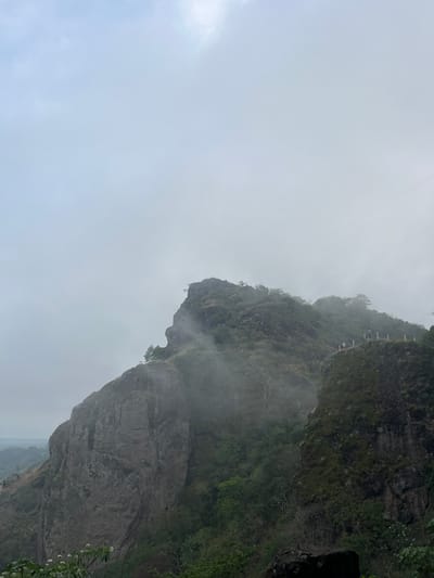 The image depicts a mountainous terrain under a cloudy sky, possibly a cliffside. The vegetation consists of greenery. A trail or path with white railings can be seen along the mountain, with figures that may be people on the path. The scene is Planes de Renderos, El Salvador.