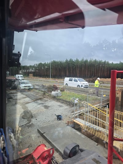 The image captures a construction scene, possibly near Wałcz, Poland, characterized by overcast skies and a damp environment. In the foreground, a red piece of heavy machinery is visible, partially obscuring the view. The mid-ground shows a construction site with numerous piles of rebar, a white car, and a white van on a road. A person wearing a high-visibility yellow jacket and a white hard hat is standing on a raised platform next to the road, looking at a device in their hands. The background features a dense forest of pine trees and a highway under construction. The overall impression is one of active construction work in a semi-rural, possibly forested area.