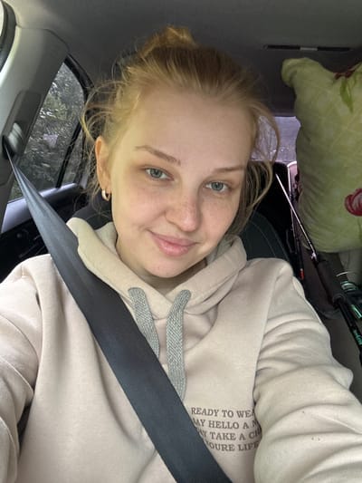 A young woman with blonde hair pulled up in a bun and a light pink hooded sweatshirt is in the driver's seat of a car. She is wearing a seatbelt across her chest. In the background, there is a green pillow with a floral pattern and two fishing rods. The scene appears to be set in Anteplevo (Okhotinskoe S/P), Russia.