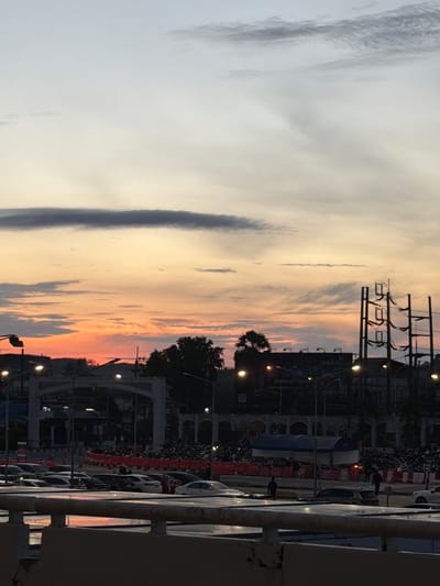 This is a vertical, eye-level shot of a parking lot at dusk, possibly in Mai Khao, Thailand, with a sunset in the background. The sky transitions from a light blue at the top to soft oranges and yellows near the horizon. Several rows of parked cars are visible in the midground, with their headlights on. A crowd of people is gathered behind orange and white barricades, suggesting an event or gathering. In the foreground, a concrete barrier runs horizontally across the bottom of the frame. Various streetlights and power poles are scattered throughout the scene, their bulbs illuminated. Buildings, including some with arched facades, are visible in the background. The overall impression is a busy urban scene at the end of the day.