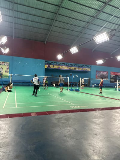 The image captures an indoor badminton court within a gymnasium, likely in Kecamatan Cisaat, Indonesia. The court surface is green with clear white demarcation lines, and a section of a darker grey concrete floor is visible to the right. Bright rectangular lights illuminate the area, suspended from the high, corrugated metal ceiling.
Several people are present on and around the court, seemingly engaged in or preparing for badminton. One person in a red sleeveless shirt and dark shorts stands on the court near the net. Another individual, wearing a light blue/grey shirt and dark shorts, is further back on the court. Closer to the foreground, two players are visible near the baseline; one is in a white and blue shirt holding a racket, and the other wears a multi-colored (green, black, yellow) shirt. A fifth person is partially seen crouching or sitting near the net posts.
The walls of the gymnasium are painted in sections, with a dark red band topped by a lighter blue/green section. A long yellow banner stretched across the red wall displays the text "SELAMAT DATANG PESERTA PARA BINTANG," which translates to "Welcome Participants, Stars." Other banners and posters are visible on the blue wall, though most text is unreadable, except for partial words like "Solidaritasprinsip." The word "PESEROS" is also visible on the green court surface. The overall atmosphere suggests an active sports facility during a game or practice session, under artificial lighting.