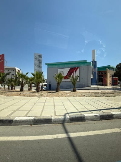 The image shows a view of a street in the Mouttagiaka Community, Cyprus, on a clear, sunny day. In the background, a tall, modern white skyscraper with a grid pattern of windows stands against a bright blue sky. To the right of the skyscraper, a low-rise building with a green roof and grey facade is visible. A large billboard on the building features a prominent image of a red stiletto heel with the words "THE DEVIL WEARS PRADA" in white text. Adjacent to this building, another structure with a red and yellow facade and a green roof has windows and an entrance. The foreground of the image is dominated by a paved area with square concrete tiles, bordered by a raised curb with black and white markings. A street with white lane markings runs in front of the paved area. Several palm trees are planted in the area between the tiled pavement and the buildings. A few cars are parked in the distance. The overall scene depicts an urban environment with commercial and residential elements.