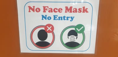 A sign stating "No Face Mask No Entry" is displayed on an orange surface, likely a wall or door. The sign has a white background with a blue border and rounded corners. At the top, the text "No Face Mask" is in red, and "No Entry" is in blue. Below the text, there are two circular icons. The left icon depicts a silhouette of a person's head and shoulders within a red circle with a red cross symbol superimposed on it, indicating prohibition. The right icon shows a similar silhouette, but this person is wearing a face mask, and there is a green checkmark in a green circle next to it, signifying approval. The image was taken in Rooppur, Bangladesh.
