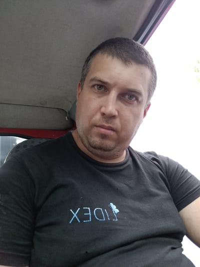 A man with short, dark hair and grey stubble is pictured in a close-up, self-taken photograph. He is wearing a black t-shirt with a light blue logo that appears to be text and a butterfly. The man is indoors, with the interior of a vehicle visible behind him, including a grey headliner and red trim. The background outside is bright white, suggesting daytime and possibly an overcast sky or a bright sun off-camera. The image was taken in Svémyslice, Czechia.