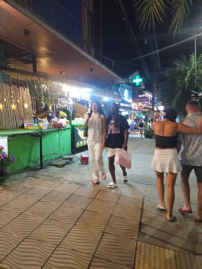The image depicts a group of people walking on a sidewalk at night. The scene appears to be a bustling street with shops and food stalls illuminated by bright lights. Palm trees are visible in the background, suggesting a tropical location. The people are dressed casually, and some are carrying bags. There are no clear indications of the weather, but the lighting suggests it is dark and likely warm. The overall mood seems relaxed and social. The visible text includes a green cross sign, possibly indicating a pharmacy, and some signage on the food stalls, though the text is largely illegible. The location is identified as Ban Klong Haeng, Thailand.