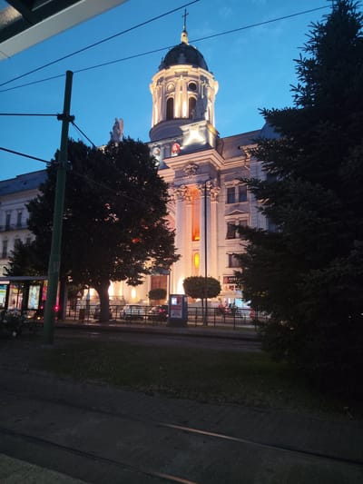The image depicts a nighttime view of a large, illuminated building, likely a church or a government building, with a prominent dome and classical architectural features. The building is lit with warm yellow lights, highlighting its columns and decorative elements. In the foreground, there are tram tracks and a grassy area. A large tree partially obscures the right side of the building. Overhead, power lines are visible, suggesting an urban environment. A tram is stopped on the left side, with advertisements visible on its windows. Several cars are parked on the street in front of the building, and a few people can be seen near the tram and on the sidewalk. The sky is a deep blue, indicating it is twilight or early night. The building in the image is the Saint Trinity Cathedral in Arad, Romania.