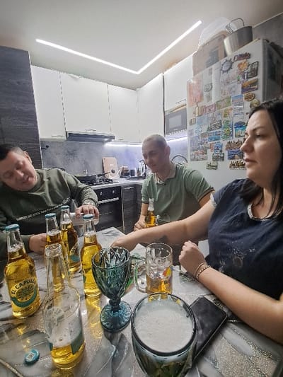 Three people are gathered around a table. Several bottles of beer labeled "El Capitan" and various glasses are on the table. One glass is full of a frothy beverage. One person is pouring liquid from a beer bottle into a glass. The setting appears to be a kitchen, with a refrigerator adorned with magnets visible in the background.
