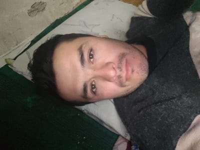 A person with dark hair and brown eyes lies on a pillow. They appear relaxed, with their head resting on the pillow and their body angled toward the right. The person is wearing a dark sweater. The background consists of a green surface, and a light-colored wall with some imperfections or patching. The lighting is somewhat soft and diffused.
