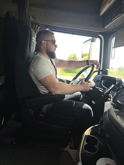 The interior of a truck cab is depicted with an adult male seated in the driver's seat. He wears glasses, a light-colored T-shirt, and dark jeans. He has a beard and short hair. He is holding the steering wheel. The dashboard and interior fixtures of the truck are visible, including the cup holders and seatbelt. Through the windshield, a rural scene is visible with fields and trees. The truck's seat has a "DAF" logo on it. The location is not identifiable as Mouscron, Belgium, based on the visual information provided.