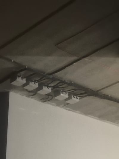 The image shows a close-up, low-angle view of a concrete ceiling with electrical conduits and junction boxes installed. Five white junction boxes are visible, spaced evenly and mounted on the underside of the ceiling. Black electrical conduits run along the ceiling, connecting to these boxes. The ceiling itself appears to be constructed of raw concrete panels, with visible seams and texture. The lighting in the scene is dim, casting shadows from the junction boxes and conduits onto the concrete surface. The overall impression is of exposed infrastructure, possibly in a building under construction or with an industrial aesthetic. The specific location in Zima Juzhnaja, Russia, is not visually ascertainable from the image content.