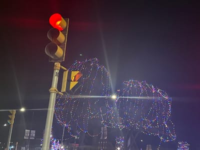 The image depicts a nighttime street scene, centered on a traffic light and an illuminated holiday display. The primary subject is a vertical traffic light on the left, with its top signal illuminated in red, indicating a stop. The two signal housings below the red light are dark. The traffic light is attached to a light-colored pole, which clearly shows the number "10" in black text.
To the right and in the background, a large, abstract shape formed by numerous multi-colored lights is visible. These small, bright lights appear in various hues including red, green, blue, and white, suggesting a festive or holiday decoration, possibly strung on a large tree or a custom structure. Bright lens flares are present in the image, originating from the red traffic light and other bright points in the scene.
The setting is outdoors at night, likely in an urban or town environment. Faint streetlights and indistinct shapes of buildings or structures can be seen in the distant background, along with what might be banners or signs, though their details are not discernible. No people are visible in the image. The visible text is the number "10" on the traffic light pole.